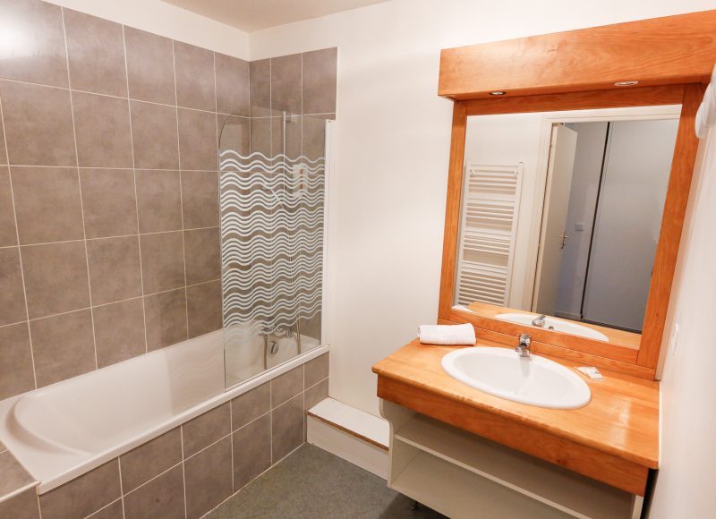 Salle de bain avec baignoire, miroir encastré dans du bois et lavabo. Sèche-serviette.